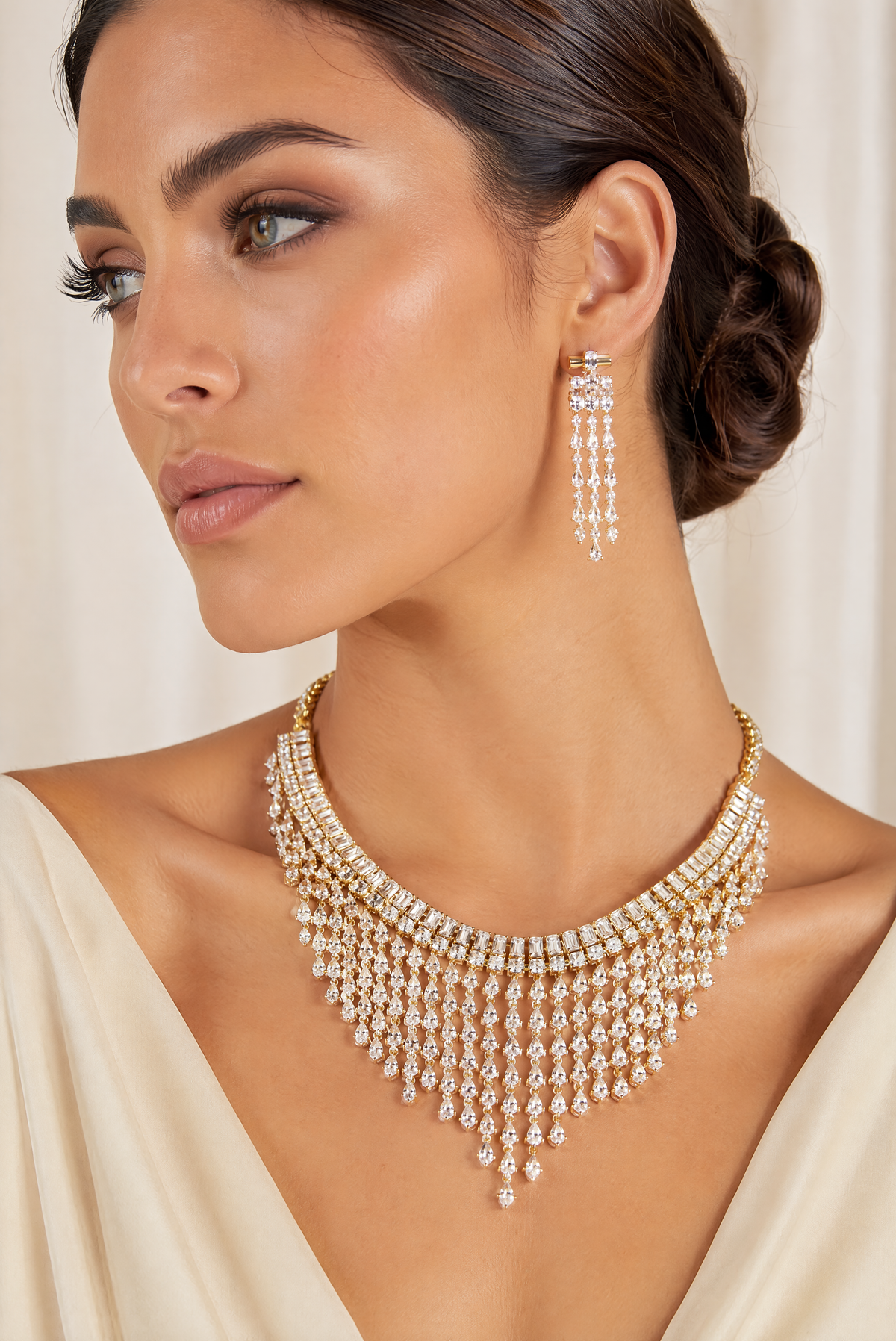 A woman wearing a gold-toned multi-strand choker necklace with dangling clear stones and matching drop earrings, shown in a close-up portrait against a soft cream background.