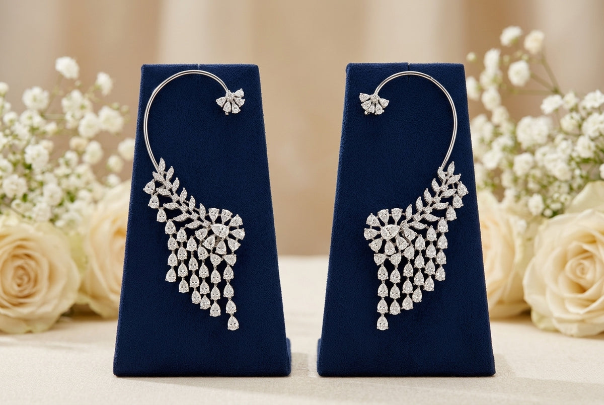 A pair of silver hoop earrings with teardrop-shaped diamond clusters, displayed on dark blue velvet stands with cream roses and baby's breath in the background.