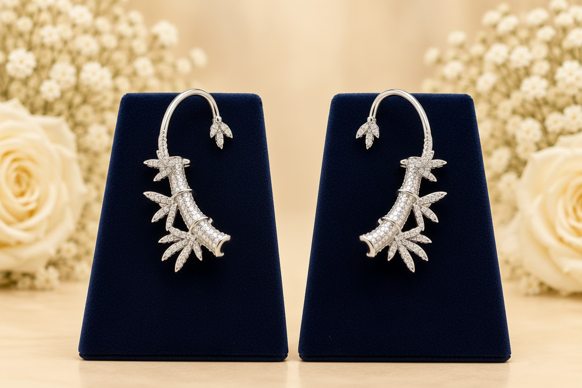 A pair of gold hoop earrings with dangling leaf and branch motifs paved with clear stones, displayed on dark blue velvet stands against a softly blurred background of cream roses and baby's breath.