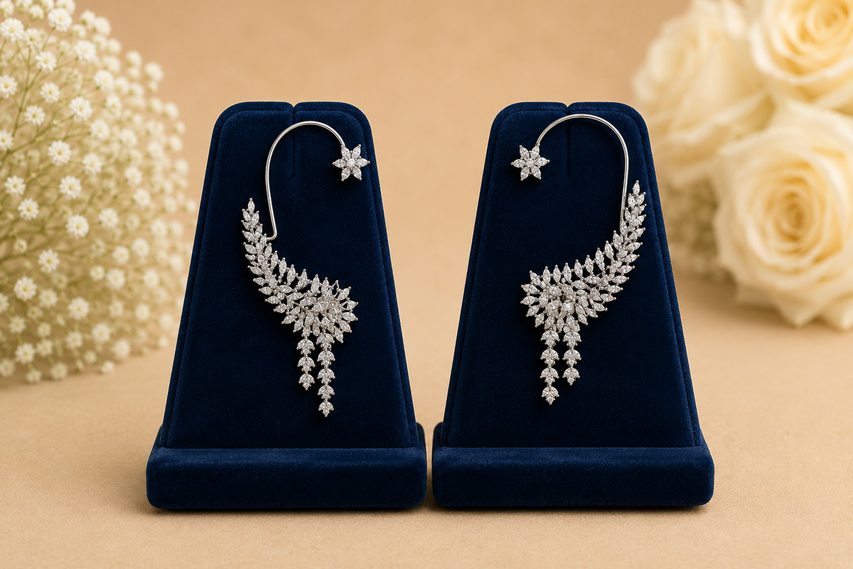 A pair of silver-toned drop earrings with pavé diamond leaf-shaped drops and starburst details, displayed on a pair of dark blue velvet jewelry stands against a beige background with blurred white flowers.