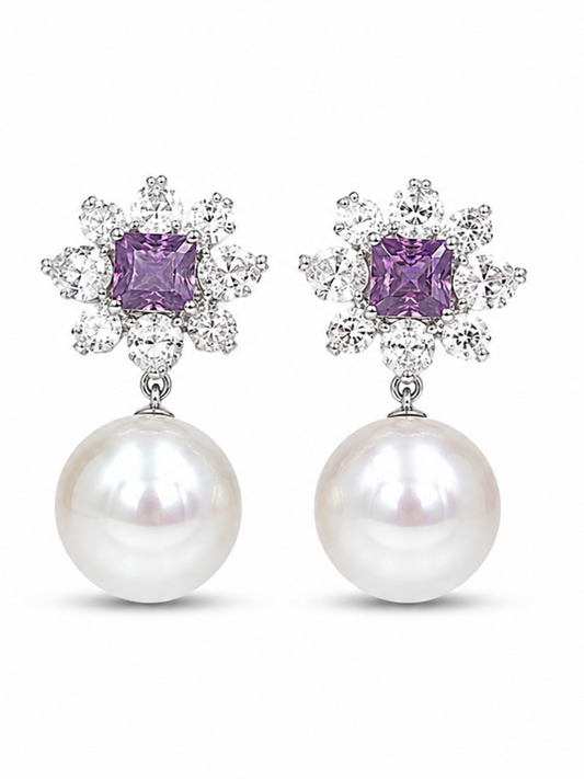 A pair of silver-tone drop earrings featuring a square-cut purple gemstone surrounded by small clear crystals, with a large white pearl hanging below, set against a plain white background.