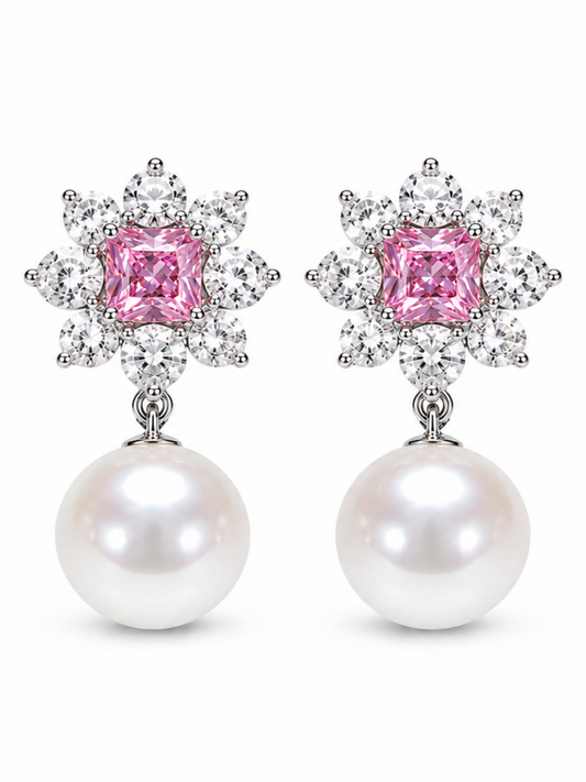 A pair of drop earrings featuring a central pink cushion-cut gemstone surrounded by a halo of small clear stones, with a large white pearl dangling below, set against a plain white background.