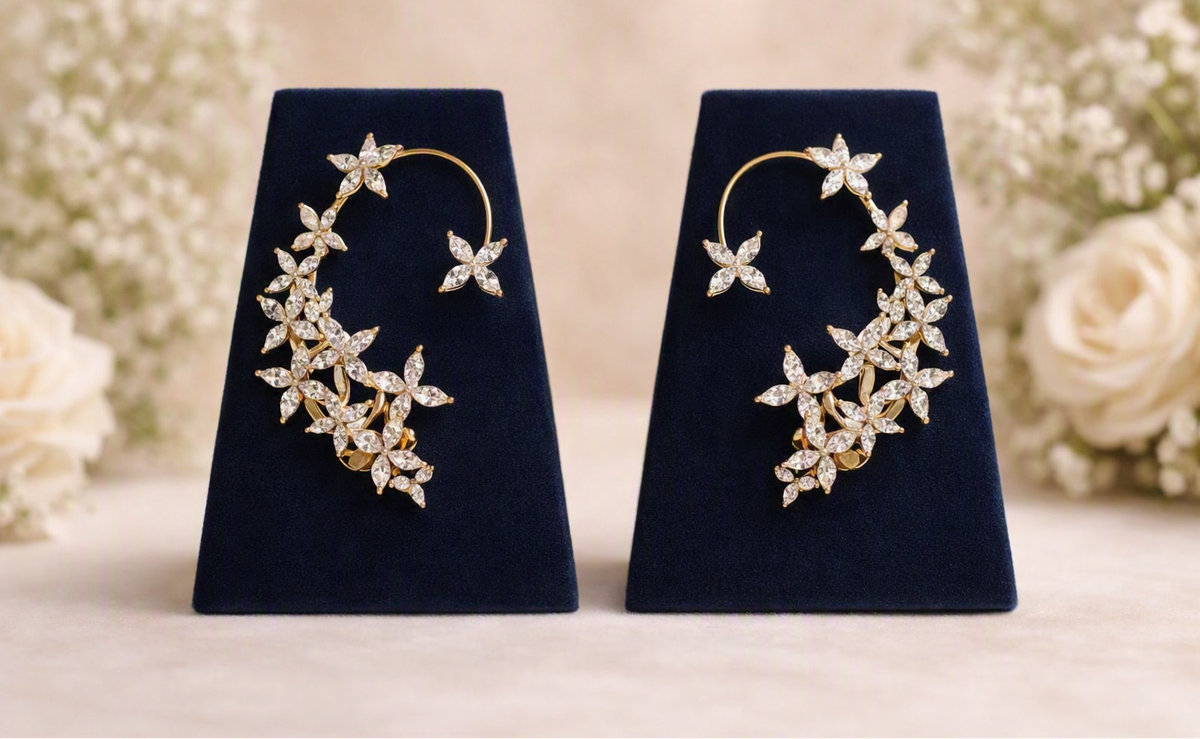 A pair of gold crescent-shaped hoop earrings with floral starburst clusters of clear crystals, displayed on dark navy velvet stands against a soft beige background with blurred white flowers. A 'Made with AI' badge is in the top right corner.