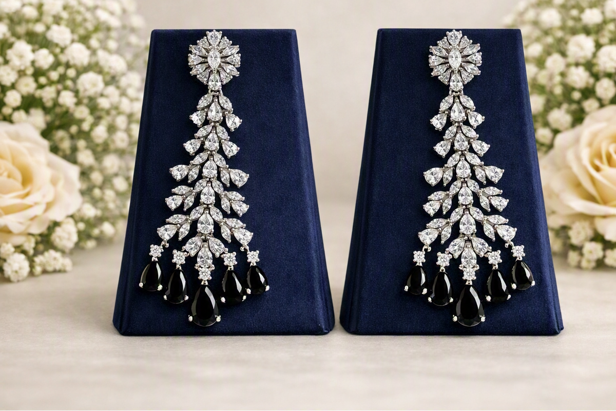 A pair of long, elaborate drop earrings displayed on dark blue velvet stands, with cream roses and baby's breath flowers in the background. The earrings feature silver-toned pavé settings with clear stones and black teardrop-shaped drops at the bottom.