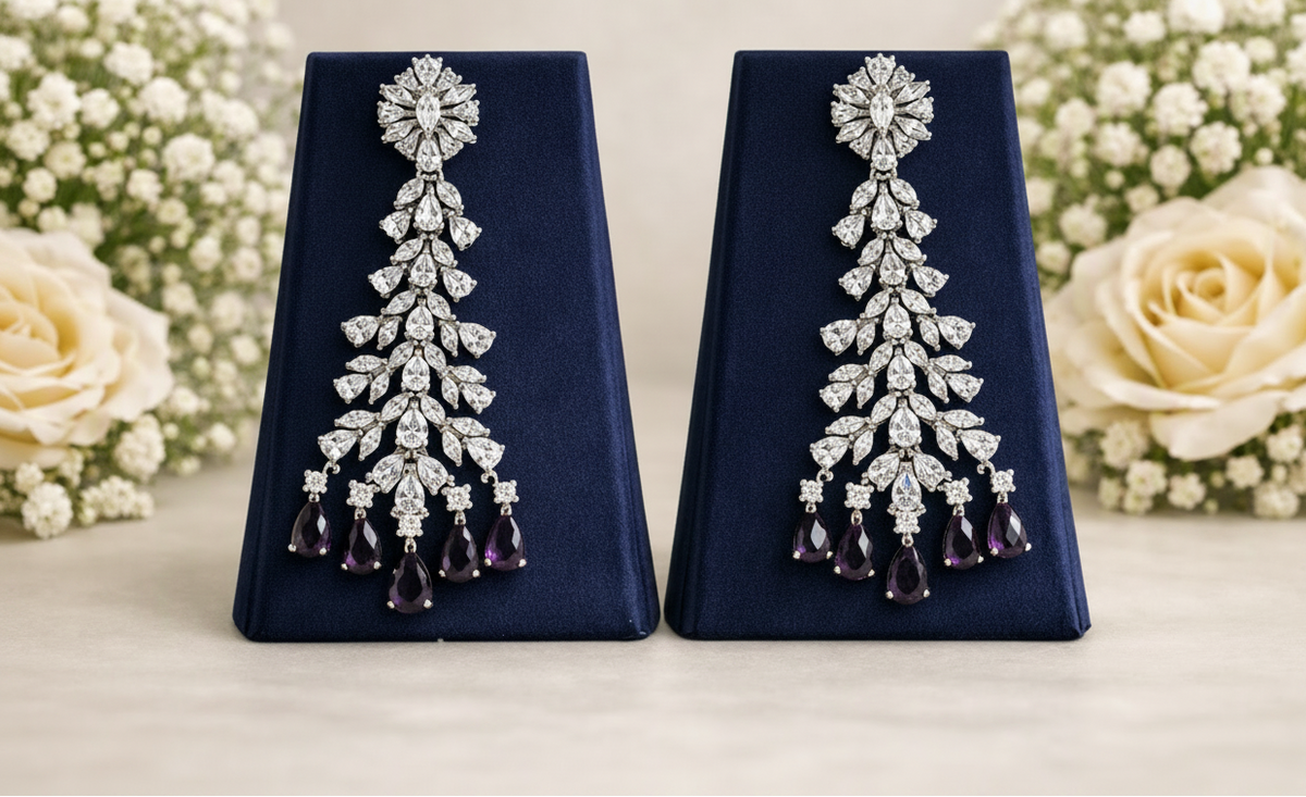 A pair of long, elaborate drop earrings displayed on dark blue velvet stands, with cream roses and baby's breath flowers in the background. The earrings feature silver-toned pavé settings with clear stones and black teardrop-shaped drops at the bottom.