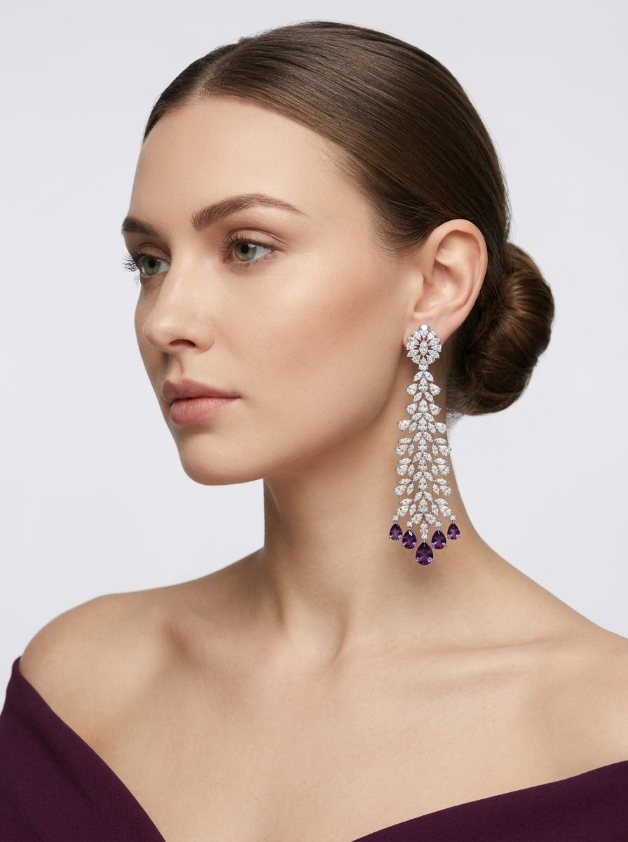 A woman with her hair in a low bun models a long, elaborate drop earring. The earring features a silver-toned setting with clear stones and dark purple crystal accents at the bottom, set against a light gray background.