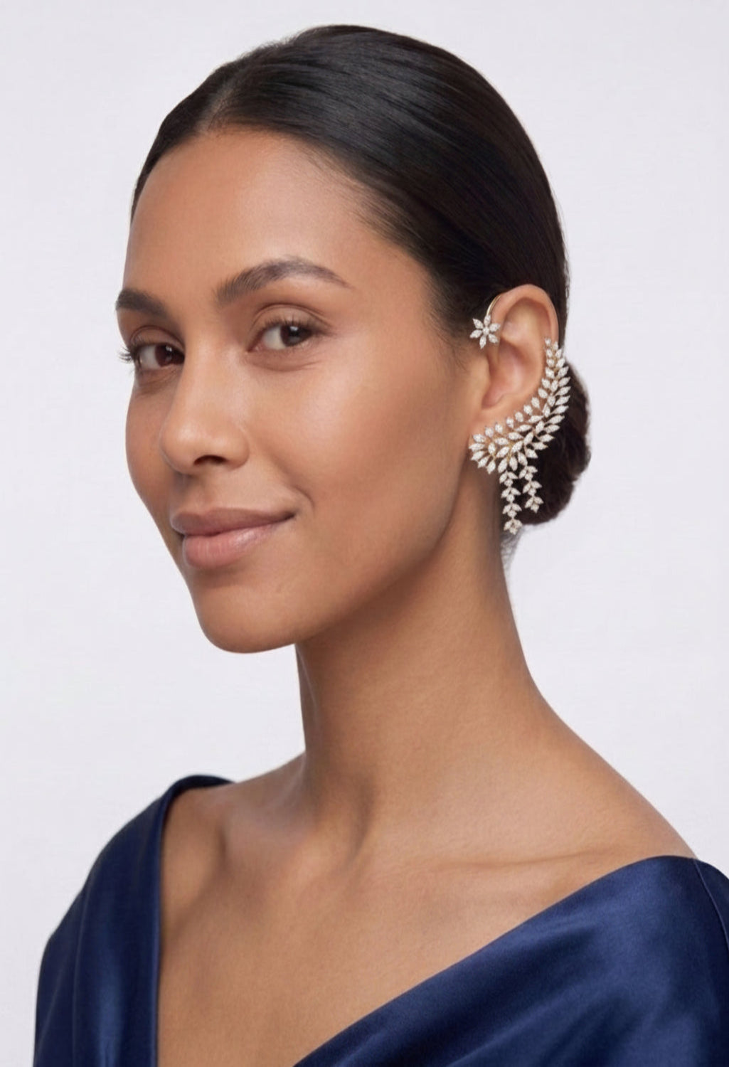 A woman with her hair pulled back wears a sparkling silver-toned leaf-shaped earring adorned with clear crystals. She is wearing a dark navy satin top against a clean, light gray studio background.