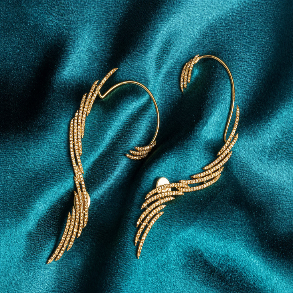 A pair of gold-toned wing-shaped ear cuffs embellished with small clear stones, displayed on a draped teal satin fabric background