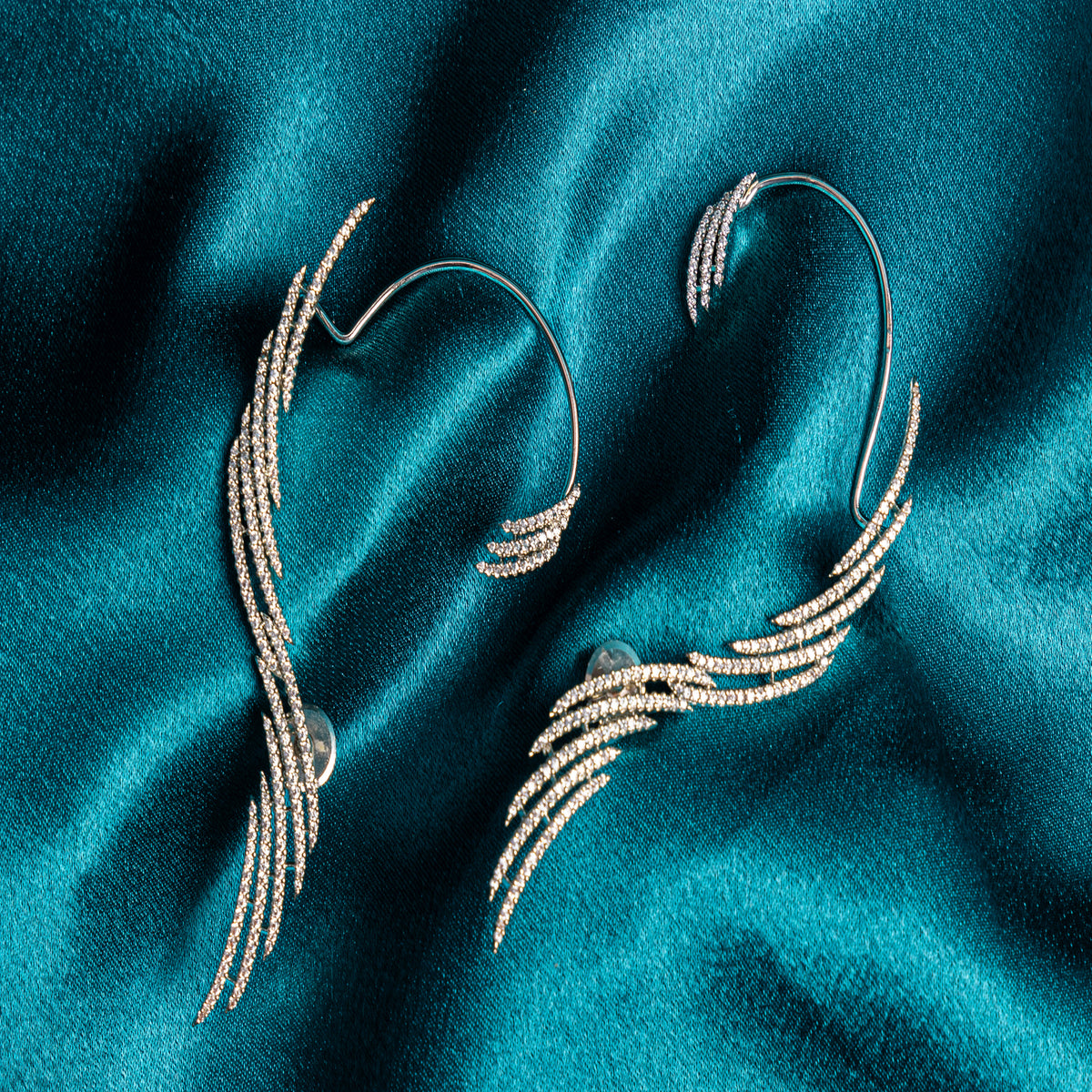 A pair of silver-toned wing-shaped ear cuffs embellished with small clear stones, displayed on a draped teal satin fabric background.
