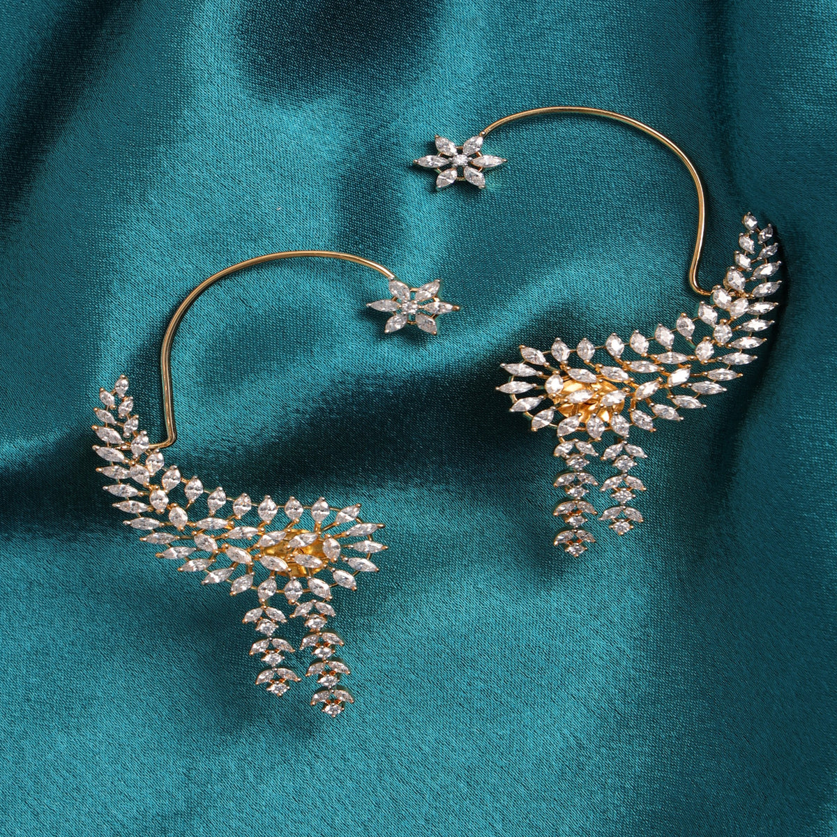A pair of gold hoop earrings with leaf-shaped clusters of clear stones and a small matching stud, displayed on a teal satin fabric background.