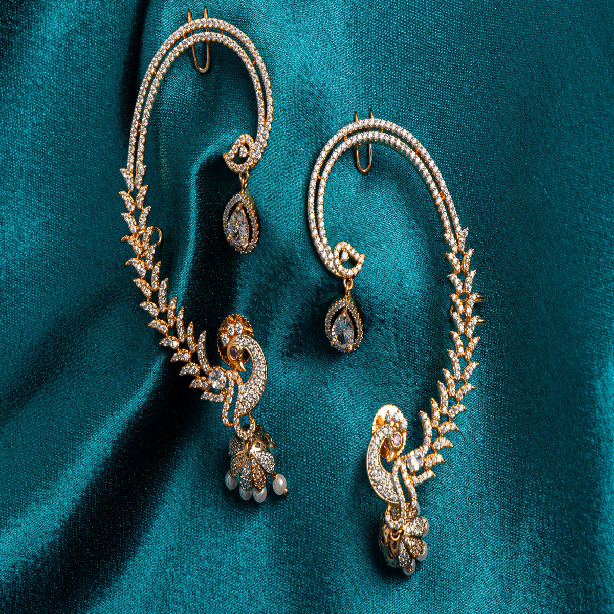A pair of ornate gold-toned ear cuffs with pavé-set stones and peacock motifs, displayed on a rich teal fabric background.