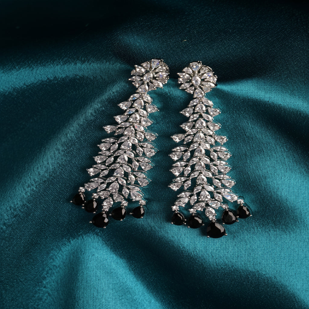 A pair of silver-toned dangle earrings adorned with clear marquise and round stones, finished with deep purple teardrop crystals at the bottom, displayed on a draped teal satin fabric.
