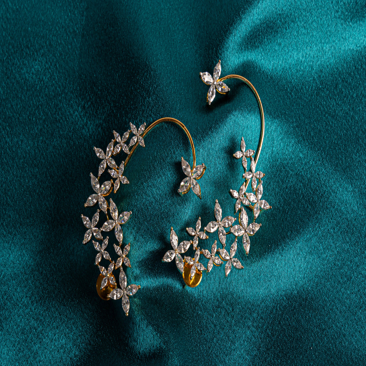 A pair of gold hoop earrings adorned with intricate floral clusters of clear pavé and round stones, displayed on a draped teal satin fabric.