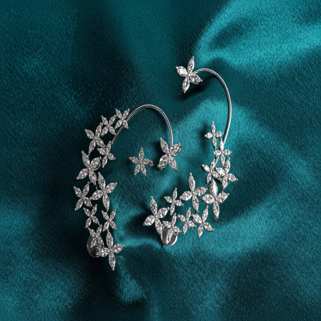 A pair of silver hoop earrings adorned with intricate floral clusters of clear pavé and round stones, displayed on a draped teal satin fabric.