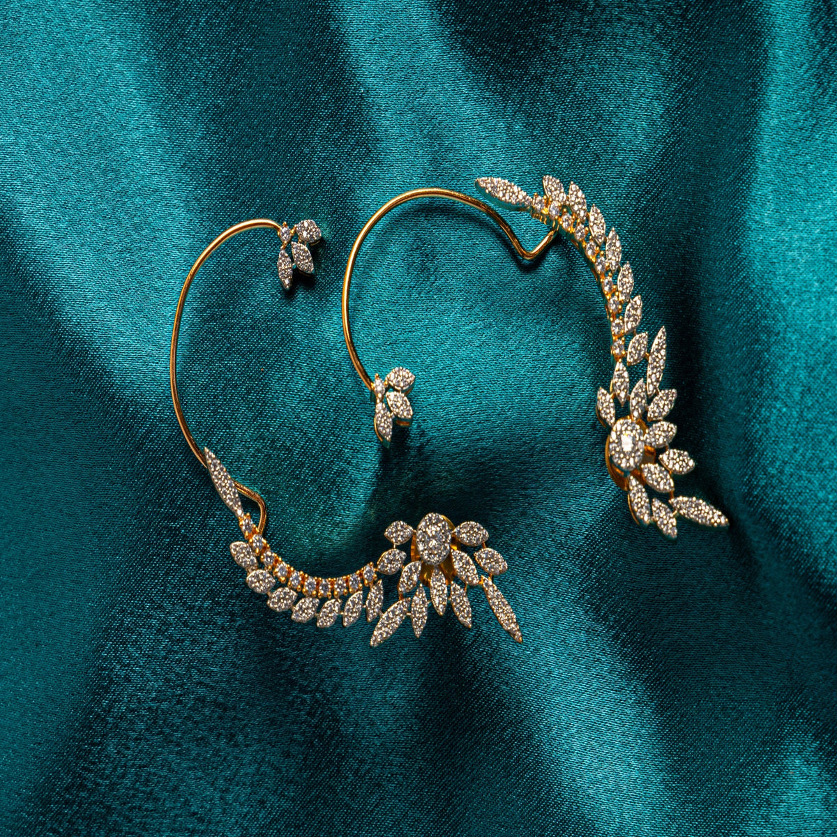 A pair of gold hoop earrings with leaf and flower motifs paved with clear stones, displayed on a textured teal fabric background.