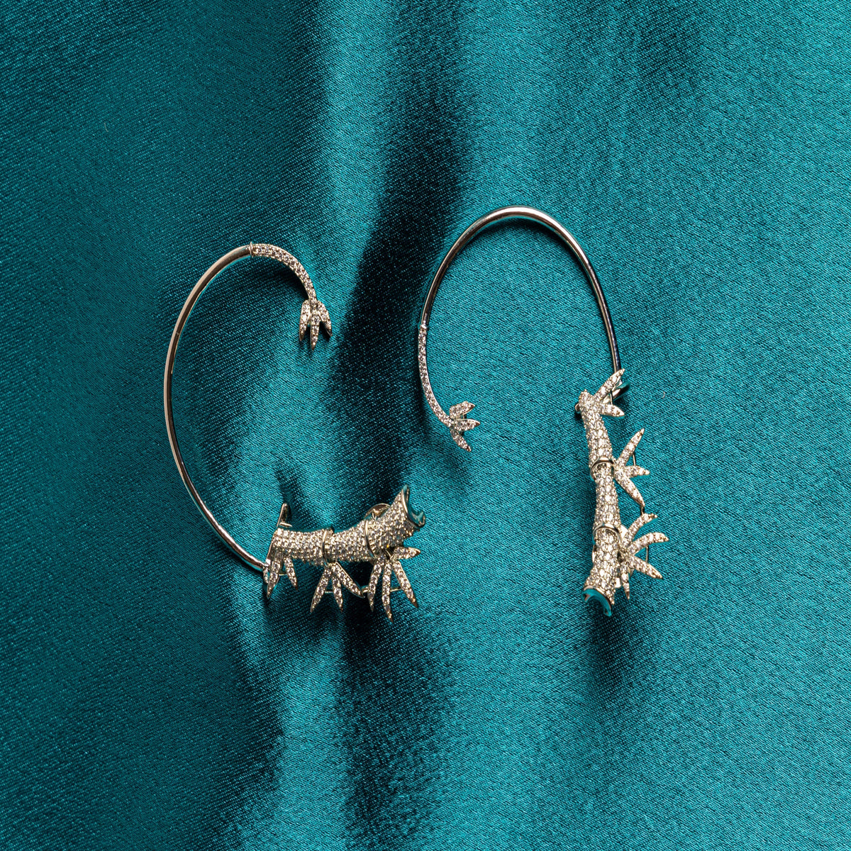 A pair of silver-toned hoop earrings with textured, coral-like antler or branch motifs at the top, displayed on a textured teal background.