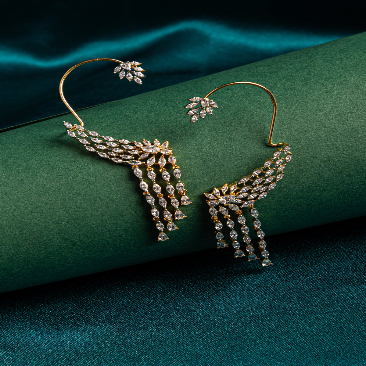 A pair of gold hoop earrings with wing-shaped clusters of clear stones and dangling fringe drops, displayed on a dark green textured surface with a matching green fabric backdrop.