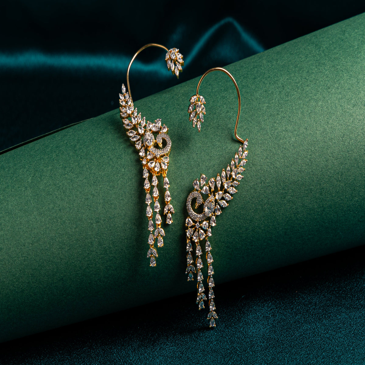 A pair of gold-toned statement earrings with pavé-set clear stones and dangling crystal drops, displayed on a dark green satin-wrapped prop against a dark teal fabric background.