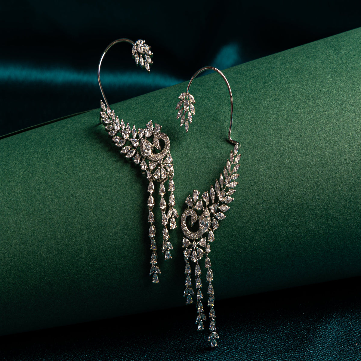 A pair of silver-toned hoop earrings with elaborate leaf and vine motifs set with clear stones, draped over a dark green velvet surface.