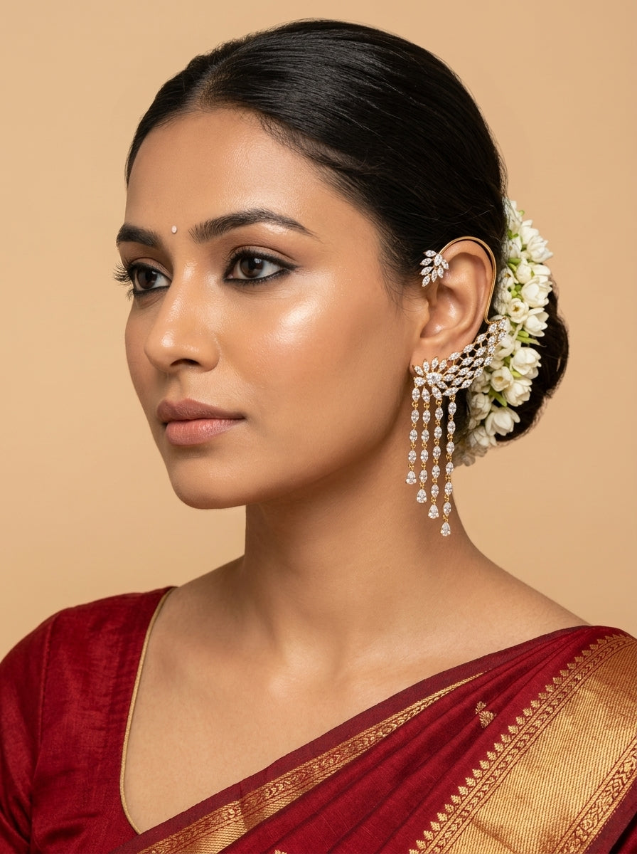 A woman in a red and gold saree wearing a silver-toned, diamond-embellished traditional Indian earring with white jasmine flowers in her hair, set against a solid beige background.
