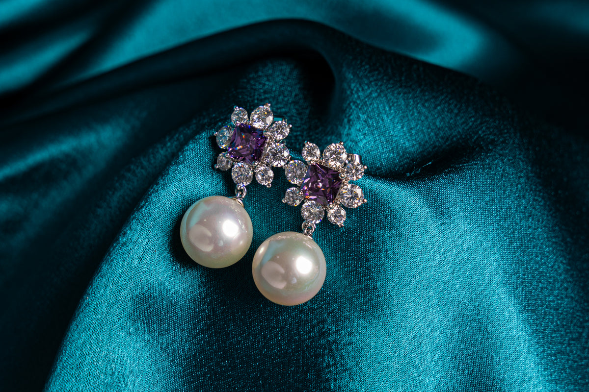 A pair of floral drop earrings with white pearls, purple gemstones, and clear crystals, displayed on a draped teal satin fabric background.