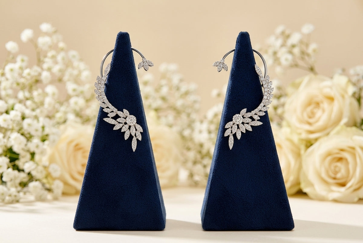A pair of silver hoop earrings with leaf-shaped crystal clusters, displayed on dark blue velvet jewelry stands against a soft beige background with blurred white flowers.