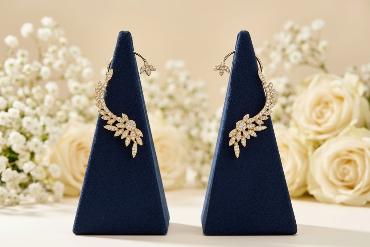 A pair of golden hoop earrings with leaf-shaped crystal clusters, displayed on dark blue velvet jewelry stands against a soft beige background with blurred white flowers.